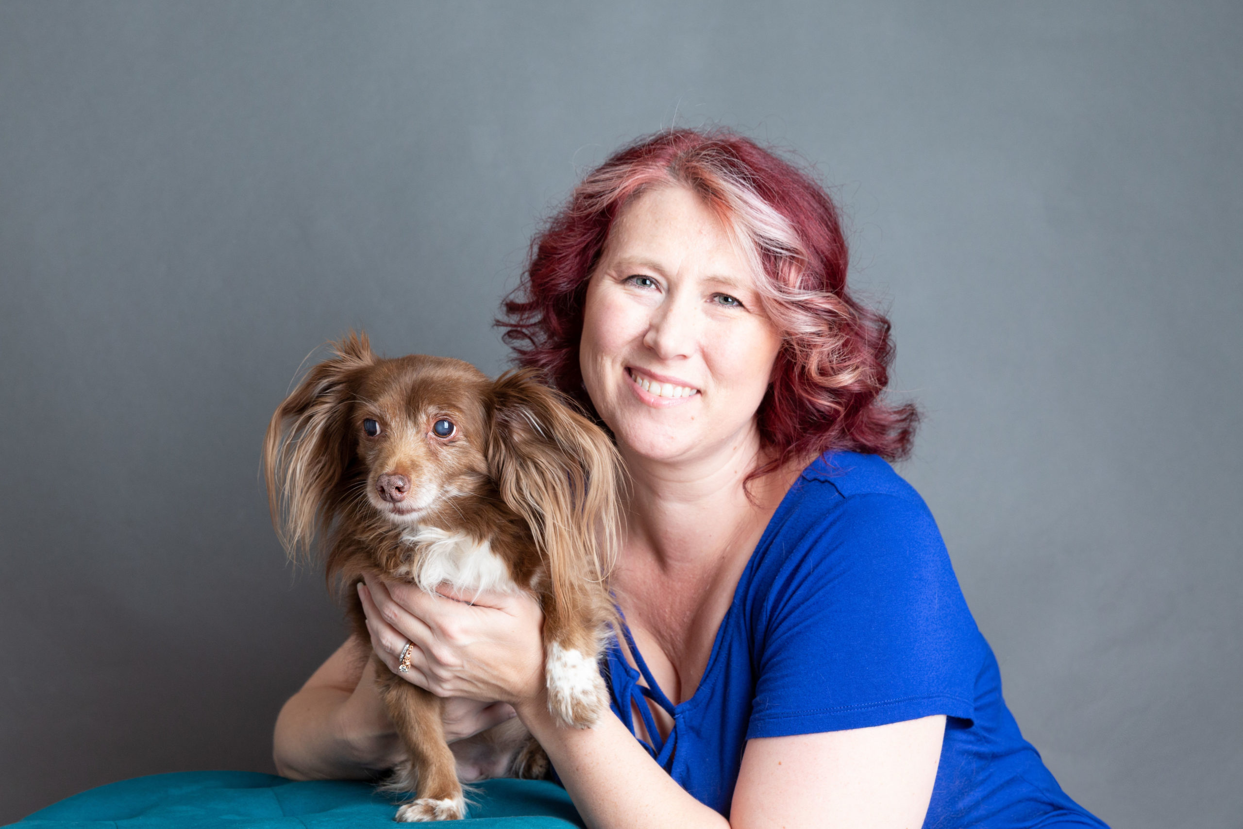 Becca in a blue shirt smiling with a brown furred pupper.